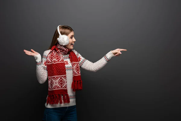 Excited woman in christmas sweater, scarf and earmuffs pointing at something, isolated on grey — Stock Photo