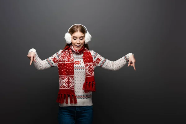 Attractive girl in christmas sweater, scarf and earmuffs pointing down, isolated on grey — Stock Photo