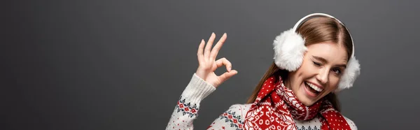 Panoramic shot of happy woman in christmas sweater, scarf and earmuffs winking and showing ok sign, isolated on grey — Stock Photo