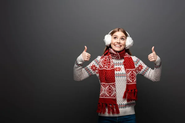 Chica feliz en jersey de Navidad, bufanda y orejeras guiñando el ojo y mostrando los pulgares hacia arriba, aislado en gris - foto de stock