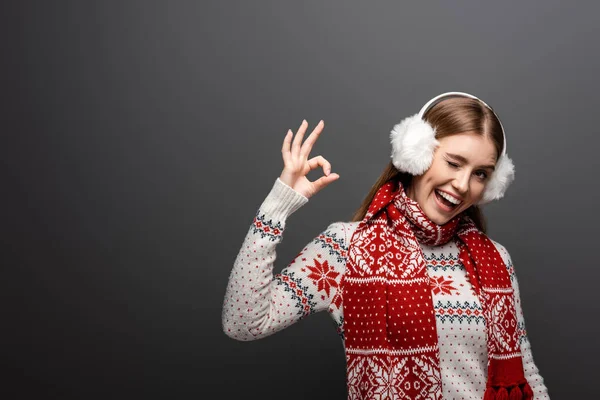 Smiling woman in christmas sweater, scarf and earmuffs winking and showing ok sign, isolated on grey — Stock Photo