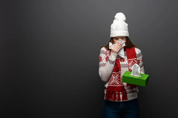 Donna malata in cappello a maglia starnutisce con tovaglioli di carta, isolata su grigio — Foto stock