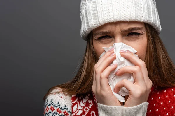 Attractive diseased woman in knitted hat with runny nose holding paper napkin, isolated on grey — Stock Photo