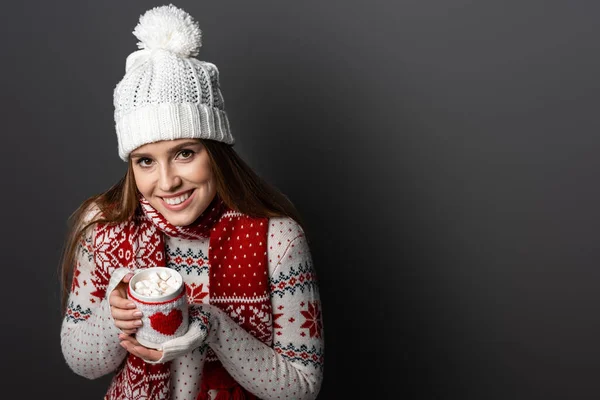 Femme souriante en chapeau et pull tenant tasse de cacao à la guimauve, isolé sur gris — Photo de stock