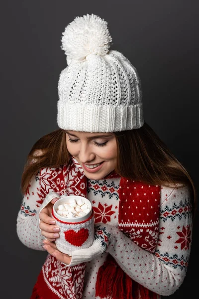 Happy woman in hat and sweater holding mug of cocoa with marshmallow, isolated on grey — Stock Photo