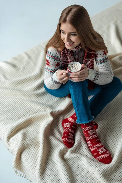 Happy woman in warm sweater holding cup of cocoa with marshmallow and sitting on blanket — Stock Photo
