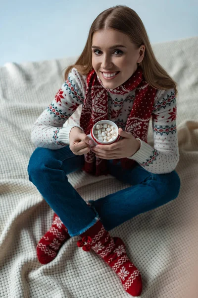 Lächelnde Frau im warmen Pullover, die eine Tasse Kakao mit Marshmallow hält und auf einer Decke sitzt — Stockfoto