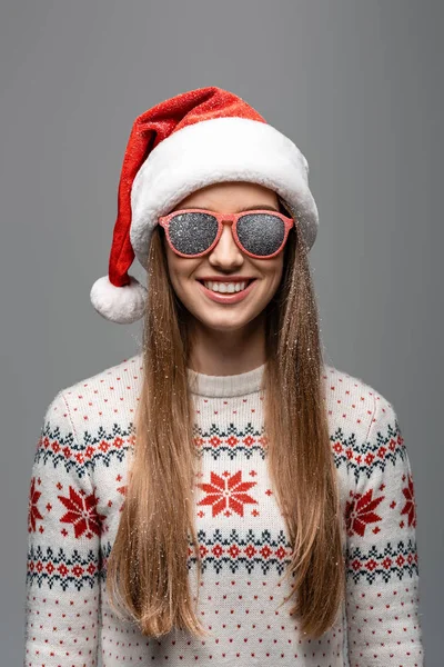 Усміхнена дівчина в різдвяному светрі, капелюсі Санти та сонцезахисних окулярах, ізольована на сірому — стокове фото