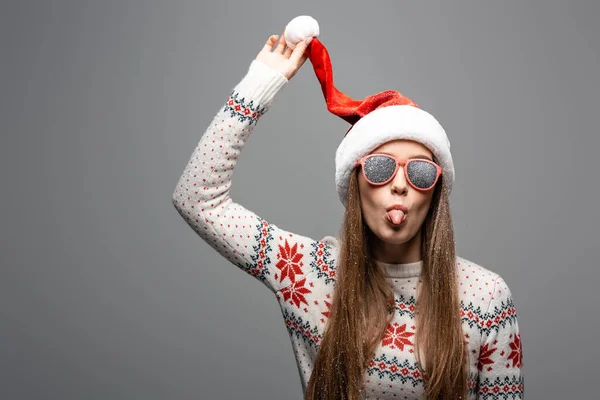 Schöne Frau im Weihnachtspullover, Weihnachtsmütze und Sonnenbrille, die die Zunge herausstreckt, isoliert auf grau — Stockfoto