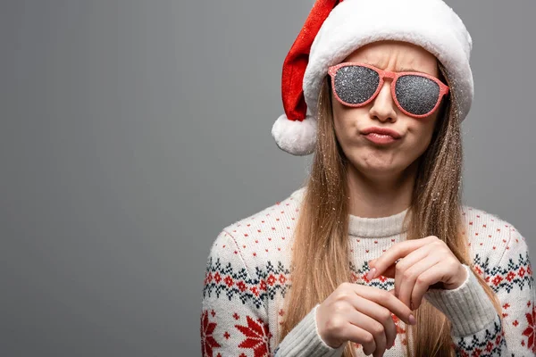 Скептически настроенная девушка в рождественском свитере, шапке Санты и солнцезащитных очках, изолированная на сером — стоковое фото
