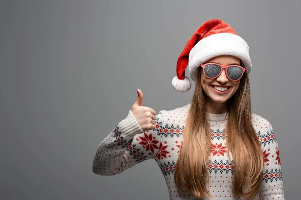 Hermosa mujer positiva en suéter de Navidad, sombrero de santa y gafas de sol que muestran el pulgar hacia arriba, aislado en gris - foto de stock