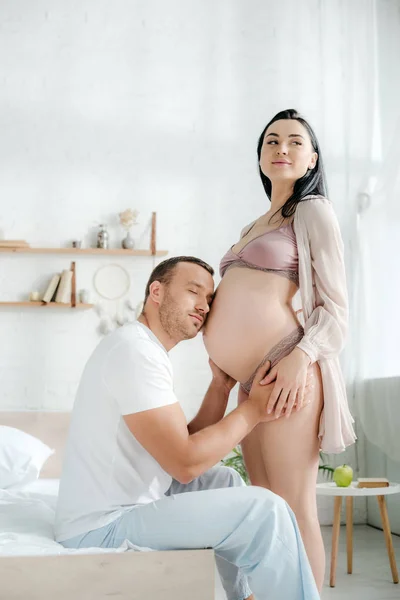 Feliz marido abrazando y escuchando vientre de su esposa embarazada feliz en la cama - foto de stock