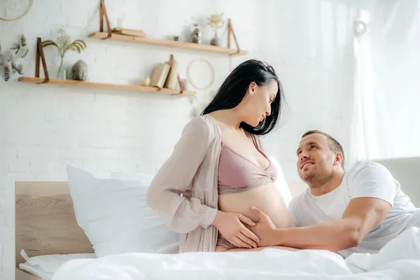 Усміхнений чоловік торкається животика своєї вагітної дружини в ліжку — стокове фото