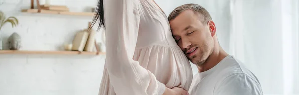 Panoramic shot of happy husband hugging belly of his pregnant wife in bedroom — Stock Photo