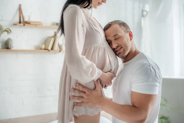 Guapo sonriente marido abrazando vientre de su esposa embarazada en el dormitorio - foto de stock