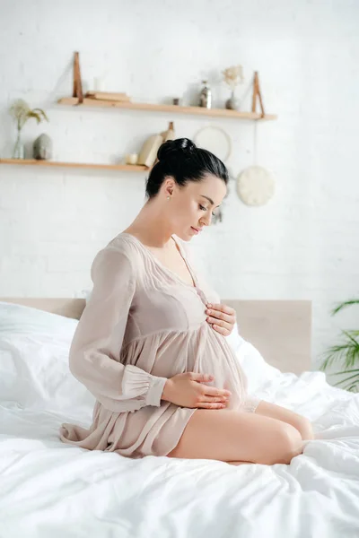 Bela mulher grávida macia em camisa de noite tocando barriga enquanto sentado na cama — Fotografia de Stock