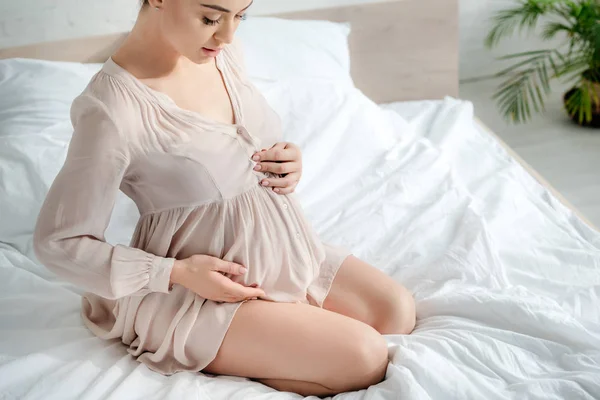 Привлекательная беременная женщина в ночной рубашке трогает животик, сидя на кровати — стоковое фото