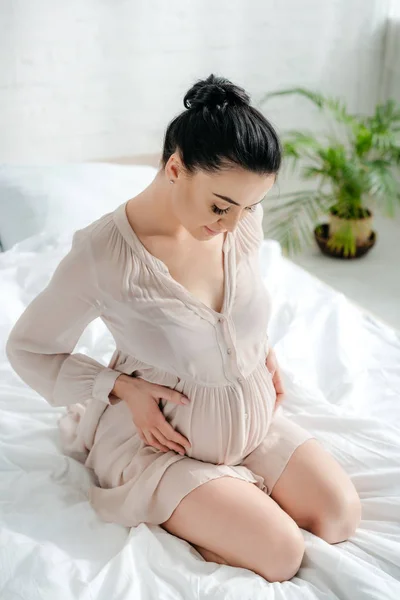 Приваблива щаслива вагітна жінка в сутінках торкається животик, сидячи на ліжку — стокове фото