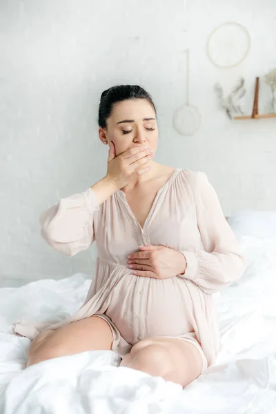 Mujer embarazada en camisón con náuseas mientras está sentada en la cama - foto de stock