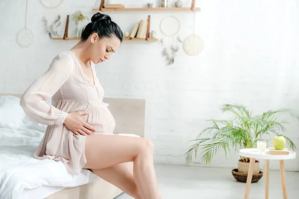 Belle femme enceinte inquiète en nuisette sentir la douleur et toucher le ventre tout en étant assis sur le lit — Photo de stock