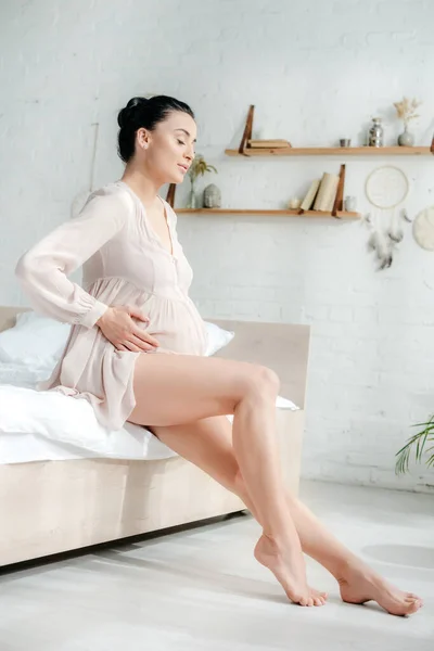 Mujer embarazada feliz en camisón tocando la barriga mientras está sentado en la cama - foto de stock