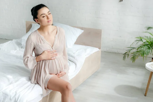 Мечтательная беременная женщина в платье трогает животик, сидя на кровати — стоковое фото