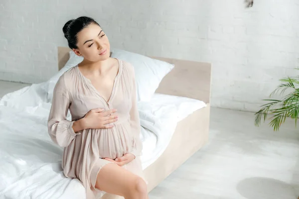 Dreamy pregnant girl in nightie touching tummy while sitting on bed — Stock Photo