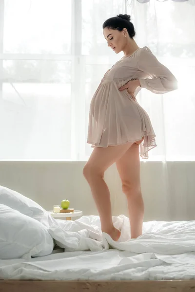 Beautiful happy pregnant woman in nightie touching tummy while standing on bed — Stock Photo