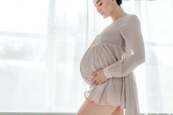Attractive happy pregnant woman in nightie touching belly in bedroom — Stock Photo