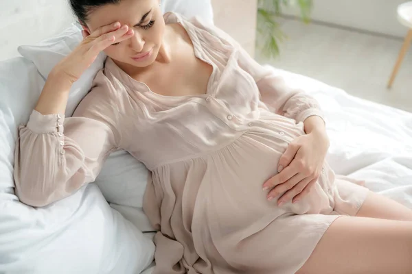 Exhausted pregnant woman in nightie having headache and touching tummy while sitting on bed — Stock Photo