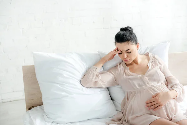 Усталая беременная женщина в ночной рубашке, у которой болит голова и трогает животик, сидя на кровати — стоковое фото