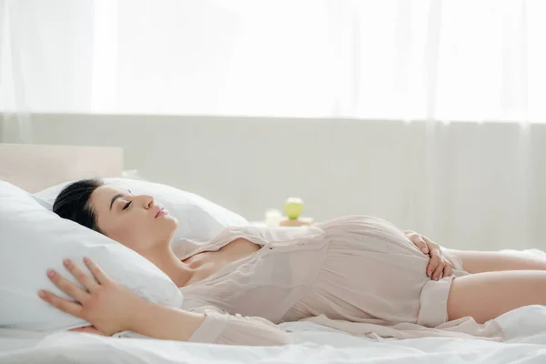 Femme enceinte en nuisette toucher le ventre tout en dormant au lit — Photo de stock