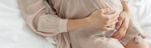 Частичный вид беременной девушки в ночной рубашке, касающейся живота во время отдыха на кровати, панорамный снимок — стоковое фото