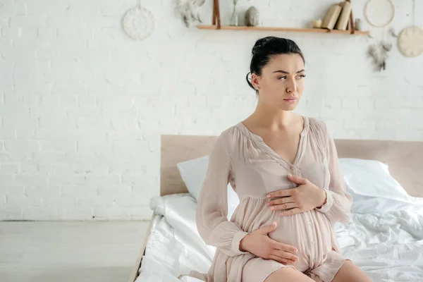 Worried pensive pregnant woman in nightie touching belly while sitting on bed — Stock Photo