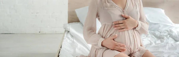 Cropped view of pregnant woman in dress touching belly while sitting on bed, panoramic shot — Stock Photo