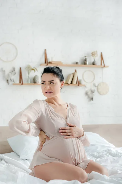 Besorgte Schwangere im Nachthemd spürt Schmerzen und berührt Bauch, während sie auf dem Bett sitzt — Stockfoto