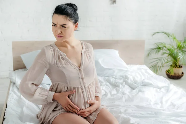 Worried pregnant woman in nightie feeling pain and touching tummy while sitting on bed — Stock Photo