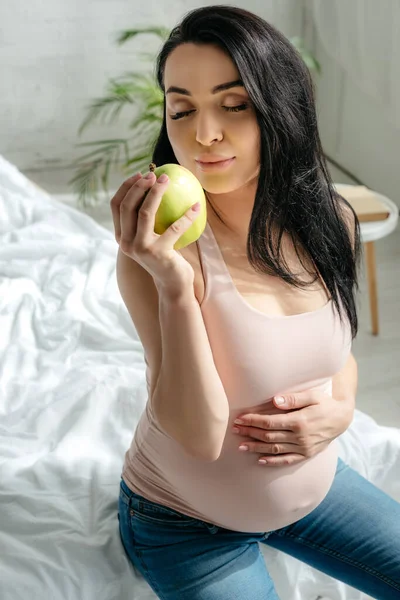 Красивая беременная женщина держит яблоко, сидя на кровати — стоковое фото