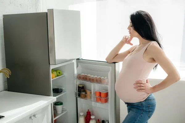 Стомлена вагітна жінка, дивлячись у відкритий холодильник на кухні — стокове фото