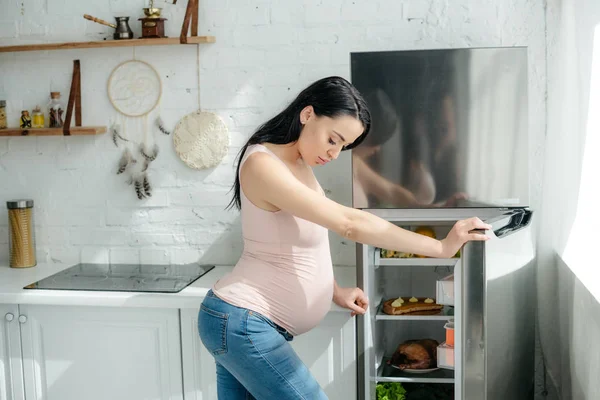 Приваблива вагітна жінка, дивлячись у відкритий холодильник на кухні — стокове фото