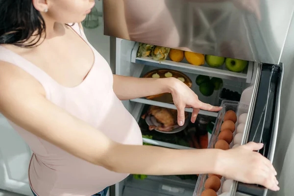 Обрезанный вид беременной девушки, указывающей в открытый холодильник на кухне — стоковое фото