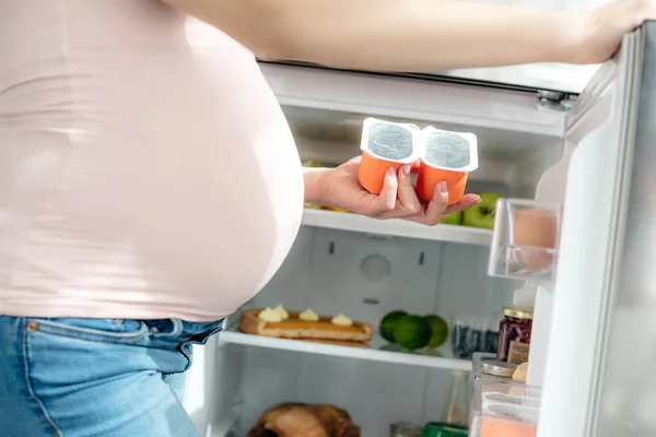 Cropped view of pregnant woman holding yogurt near opened fridge in kitchen — Stock Photo