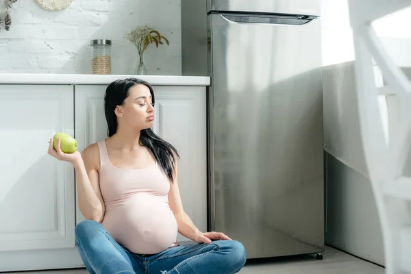 Красива вагітна жінка тримає яблуко, сидячи на підлозі на кухні біля холодильника — стокове фото