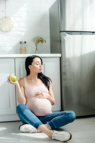 Fille enceinte tenant pomme tout en étant assis sur le sol dans la cuisine près du réfrigérateur — Photo de stock