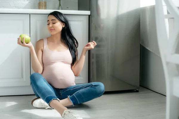 Скептична вагітна жінка тримає яблуко, сидячи на підлозі на кухні біля холодильника — стокове фото