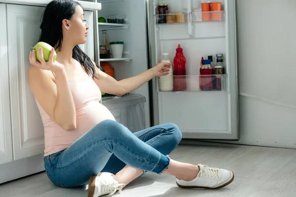 Mujer embarazada sosteniendo manzana mientras está sentada en el suelo y mirando la nevera abierta en la cocina - foto de stock