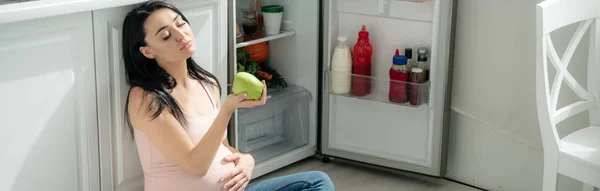 Панорамний знімок втомленої вагітної жінки, дивлячись на яблуко, сидячи на підлозі на кухні біля відкритого холодильника — стокове фото