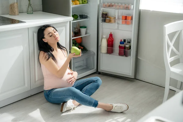 Mujer embarazada cansada sosteniendo manzana mientras está sentada en el suelo en la cocina cerca de nevera abierta - foto de stock