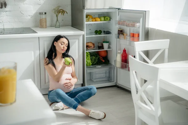 Atractiva mujer embarazada sosteniendo manzana mientras está sentado en el suelo en la cocina cerca de nevera abierta - foto de stock