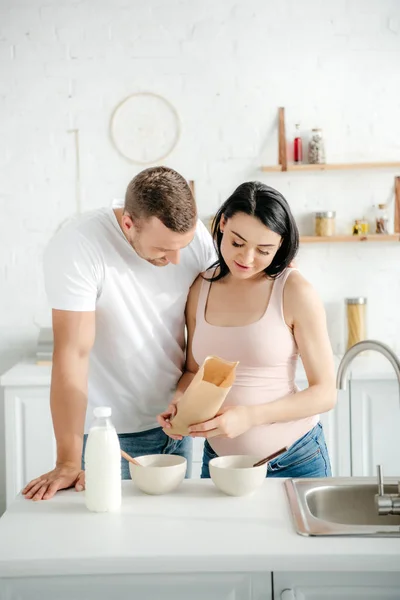 Счастливая беременная пара с зерновыми и молоком на кухне — стоковое фото